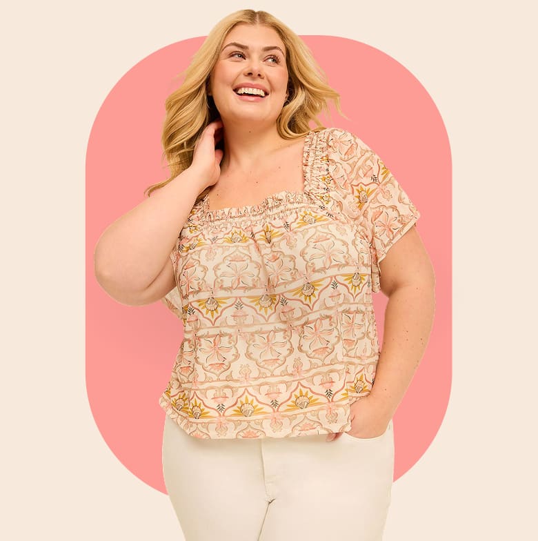 A smiling woman in a patterned blouse and white pants stands against a soft pink oval background.