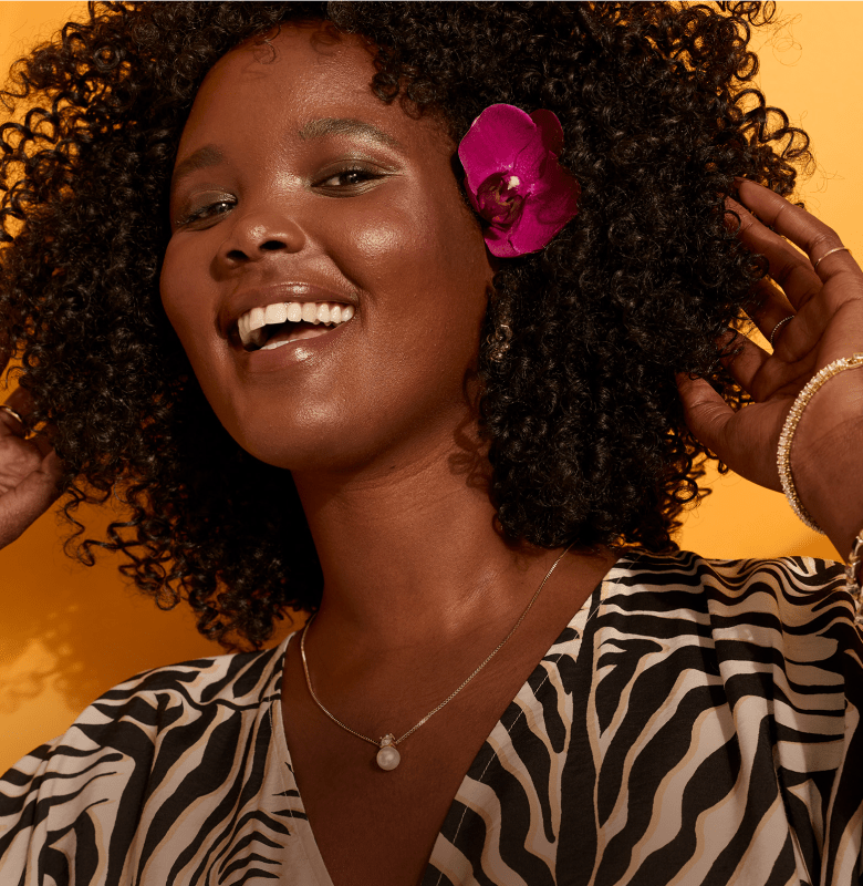 A smiling woman with curly hair adorned with a pink orchid and wearing a zebra-print top is in the foreground, with a bright orange background behind her.
