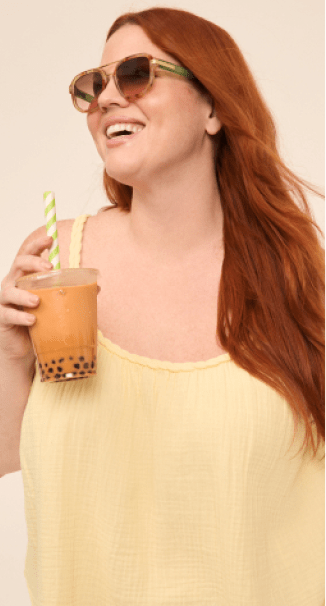 A smiling woman with red hair and sunglasses holds a bubble tea in the foreground against a plain, light-colored background.