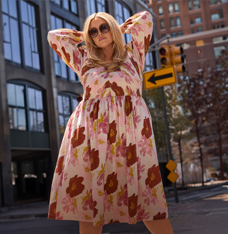 A woman in a floral dress poses with her hands behind her head in front of a city street with buildings and a traffic light.