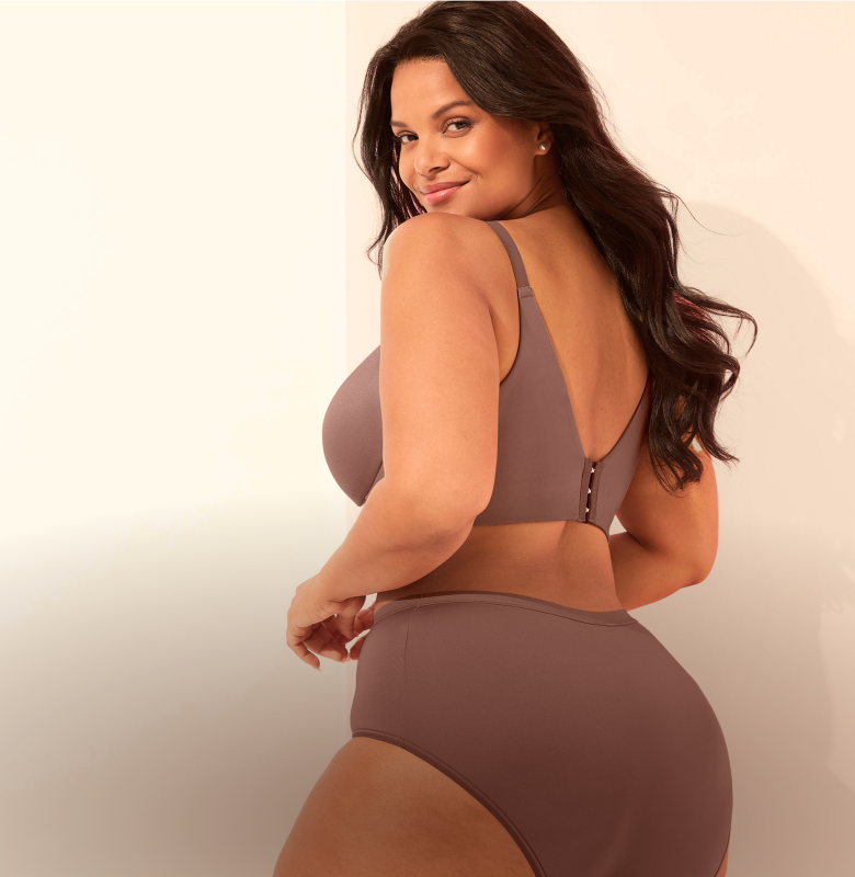 A woman in a mauve bra and high-waisted underwear poses against a plain, light-colored wall.