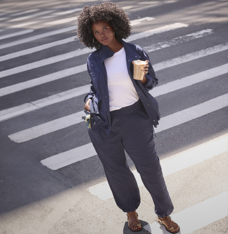 A woman with curly hair stands on a crosswalk holding a coffee, with a shadow cast across the white stripes of the street.