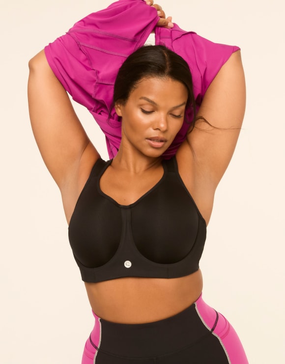 A woman in a black sports bra and pink-accented leggings pulls a magenta top over her head against a plain, light background.