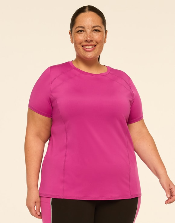 A smiling woman with freckles in a bright pink athletic shirt stands against a plain, light-colored background.