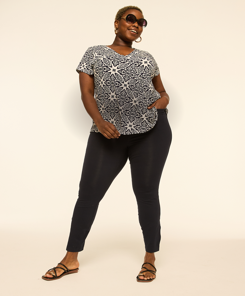 A woman in a black top and plaid pants stands in the foreground holding a black handbag, with a soft, light-colored background behind her.