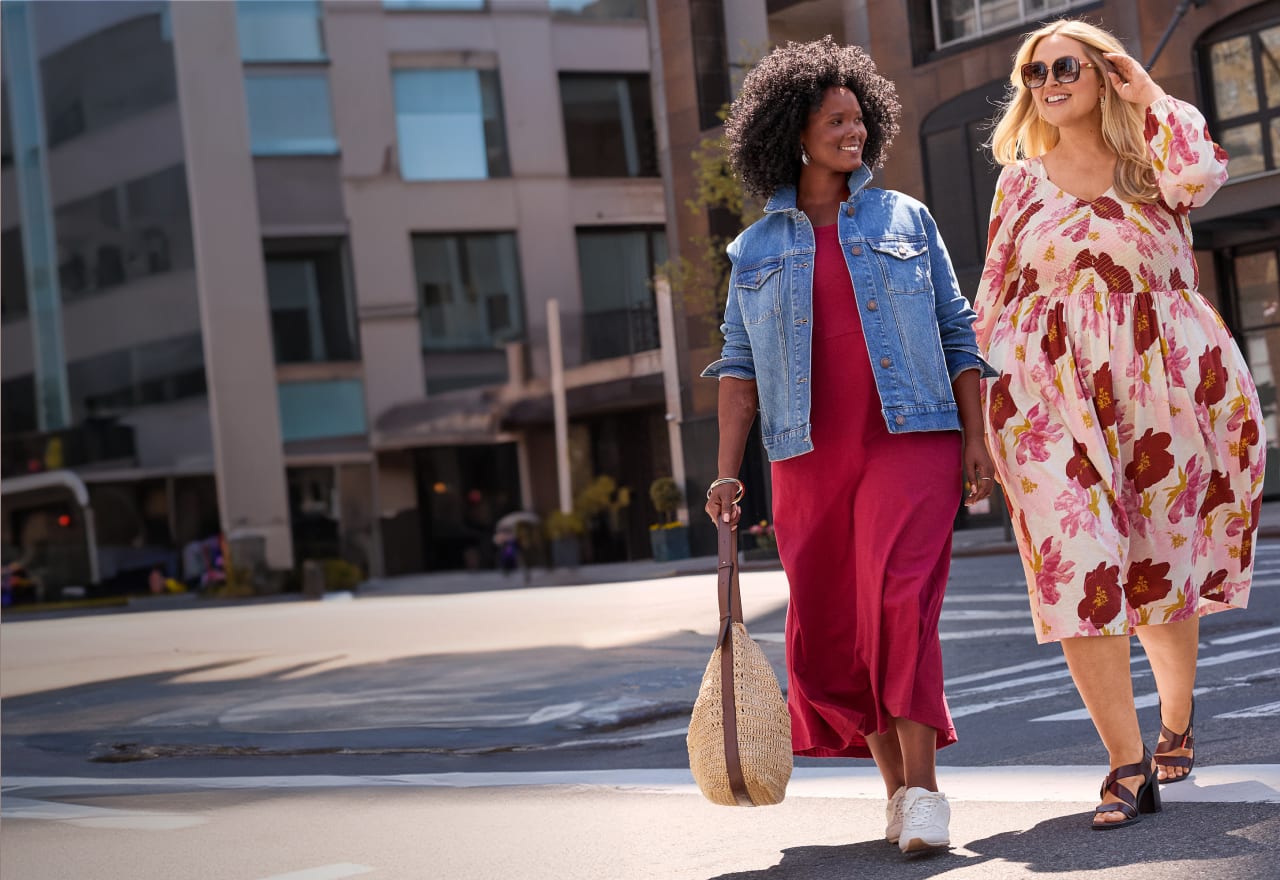 Two women walk across a city street in front of modern buildings.