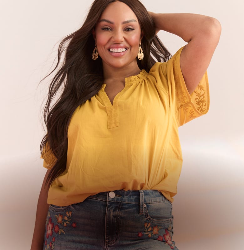 A smiling woman with long dark hair and large earrings poses in a yellow top and embroidered jeans against a soft, light background.