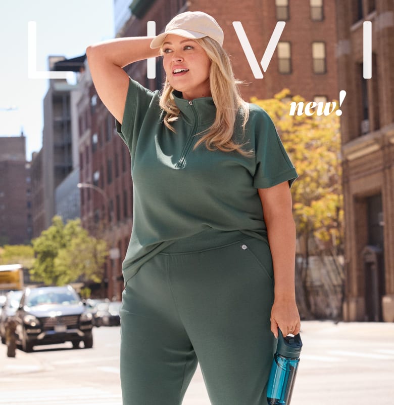 A woman in a green tracksuit and a beige cap poses with a water bottle in the foreground, while a blurred city street with cars and buildings forms the background.