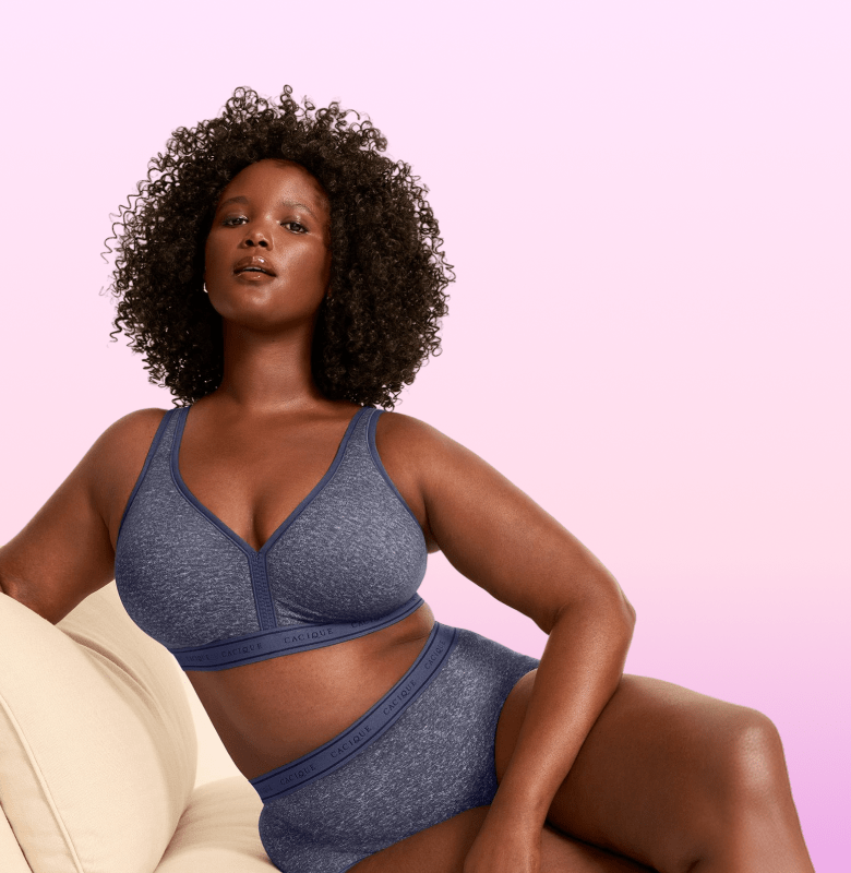 A woman with curly hair poses in a blue bra and underwear on a cream-colored couch against a pink and purple gradient background.