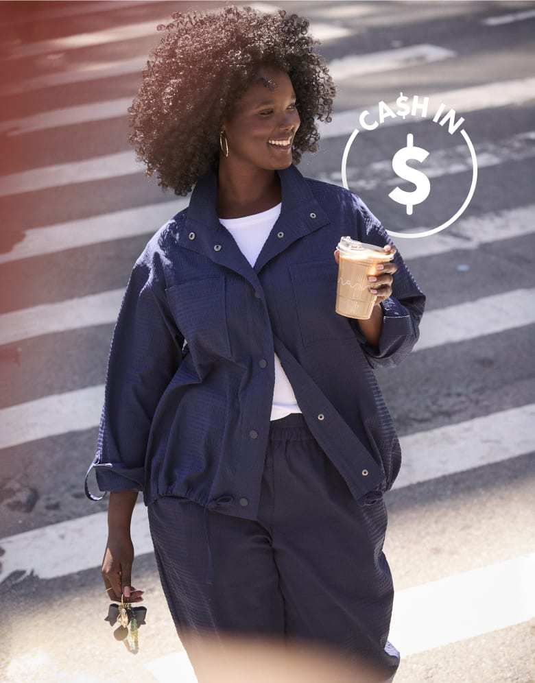 A smiling woman in a navy blue outfit walks across a crosswalk holding a drink, with a 