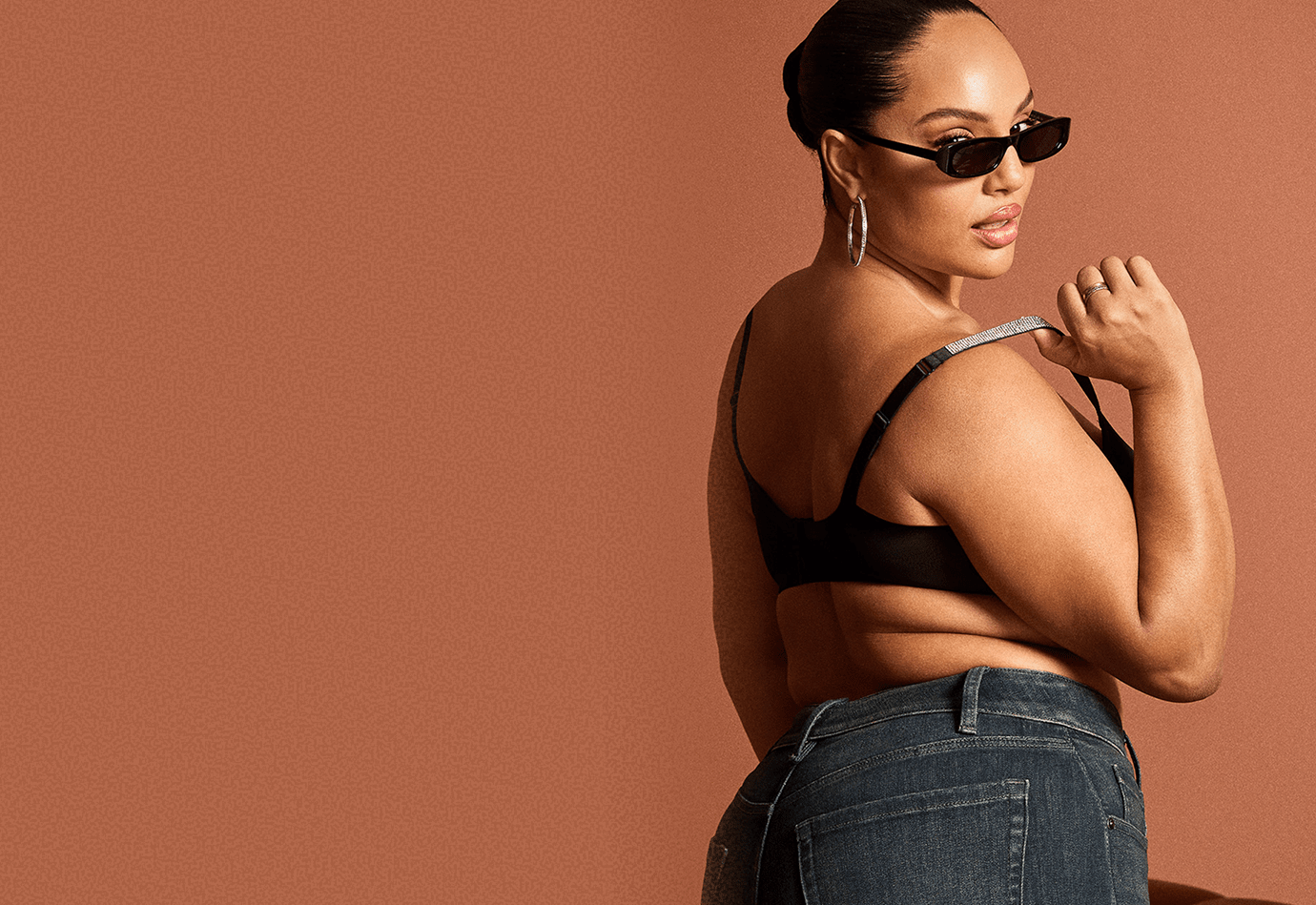 A woman in a black bra and denim jeans poses against a plain brown background.