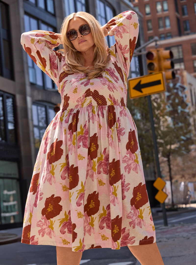 A woman in a floral dress poses with her hands behind her head in front of a city street with traffic lights and buildings.