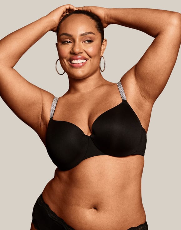 A smiling woman in a black bra with jeweled straps poses with her hands behind her head against a plain, light-colored background.