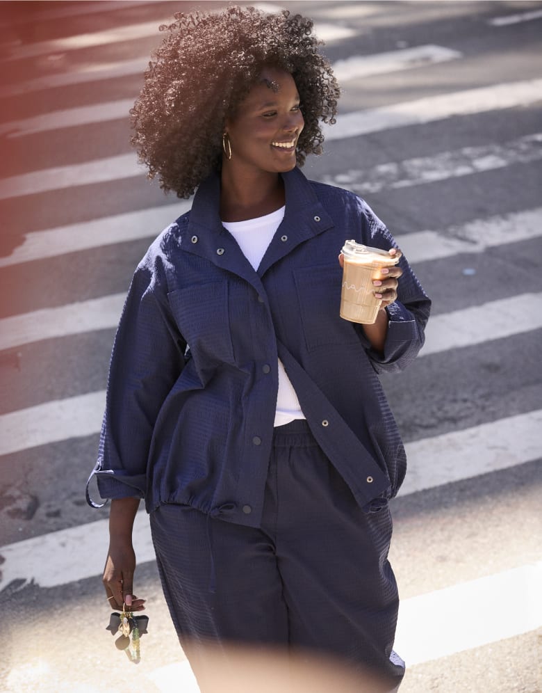 A smiling woman in a navy blue outfit walks across a crosswalk holding a drink, with a 