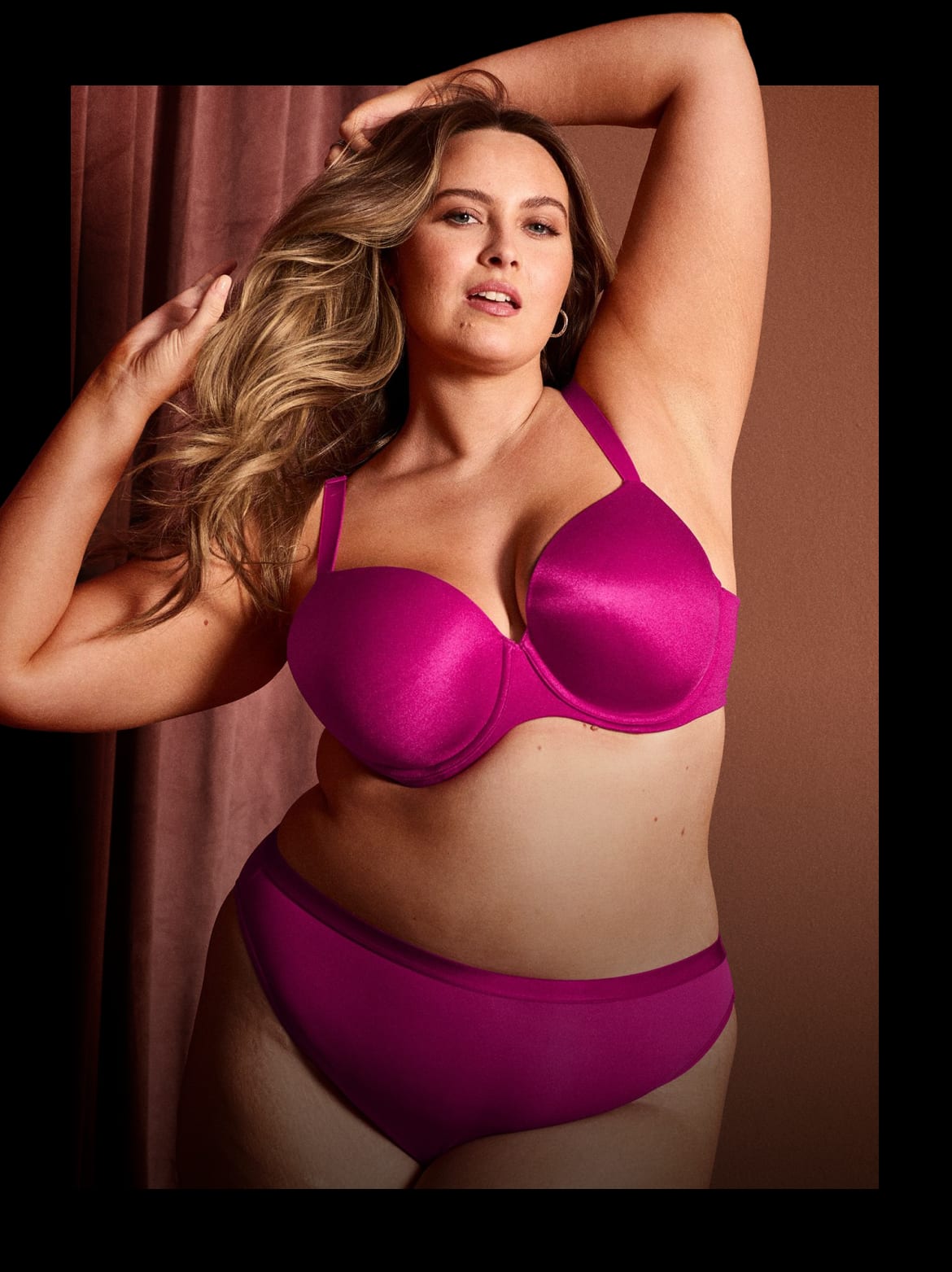 A woman in a magenta bra and underwear poses with her hand in her hair against a muted pink curtain and brown background.