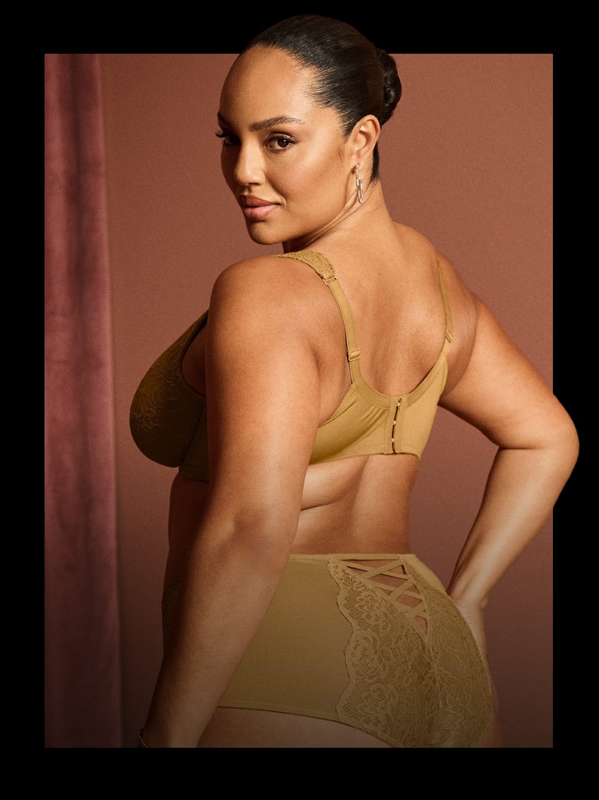 A woman in tan lingerie poses against a muted pink backdrop with a draped curtain.
