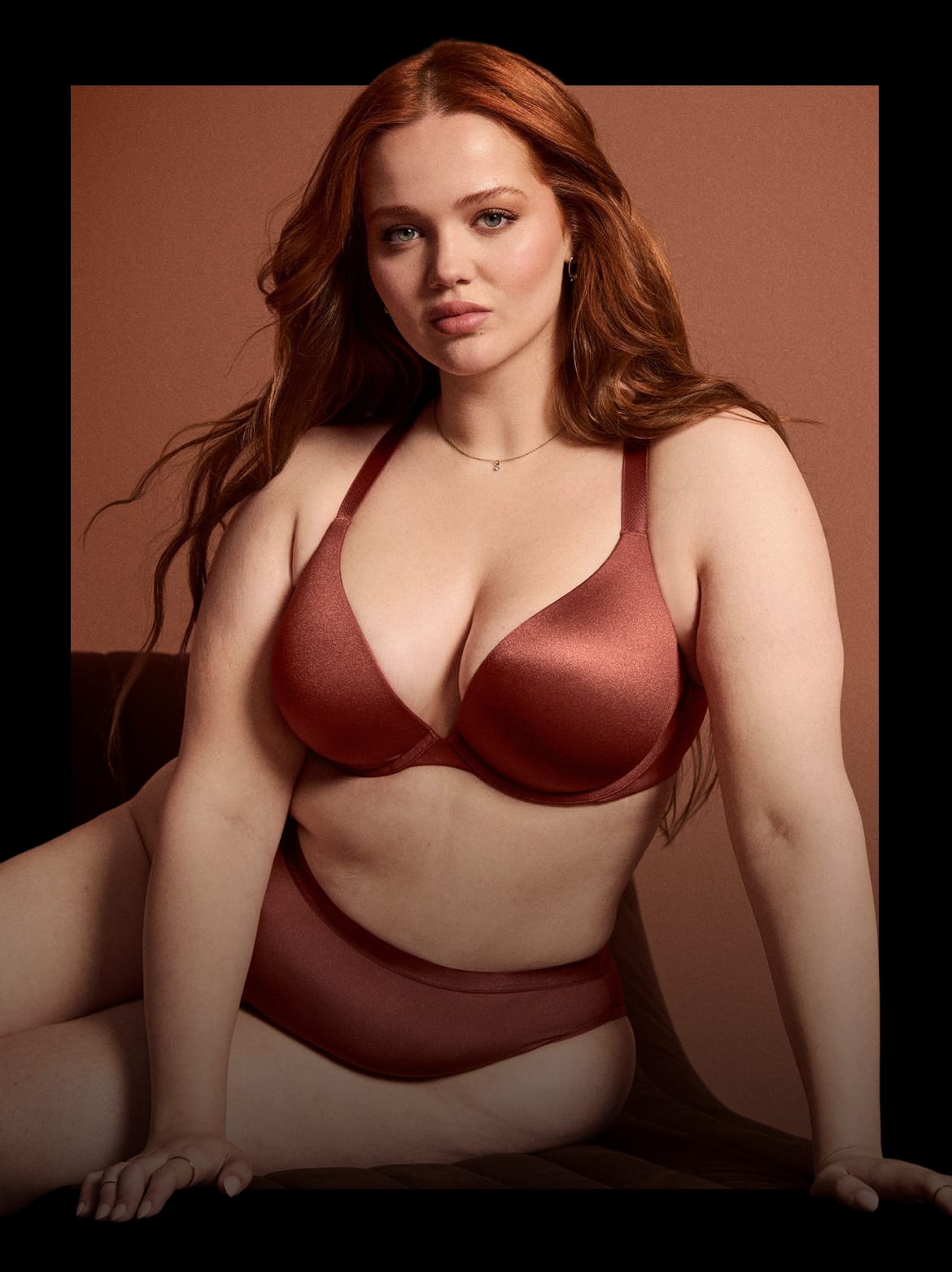A woman with long, auburn hair poses in a rust-colored bra and underwear against a warm, brown background.