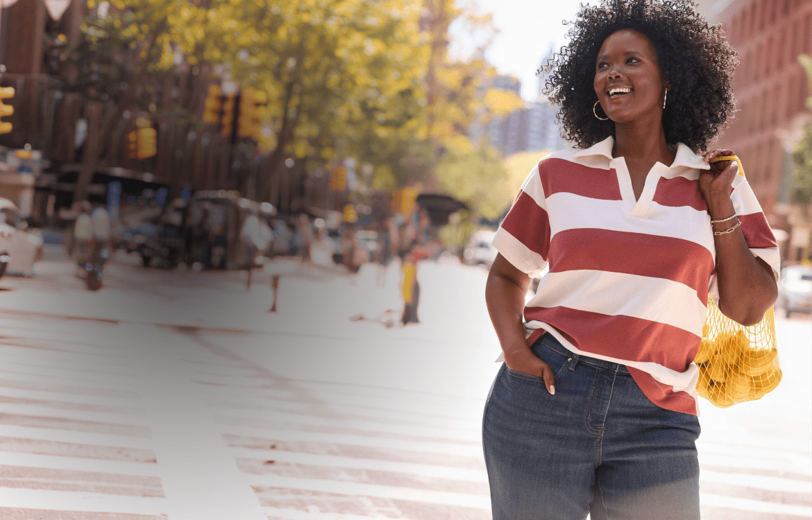 A smiling woman in a striped shirt and jeans walks across a crosswalk with a mesh bag of yellow items, while a blurred city street with cars and buildings forms the background.