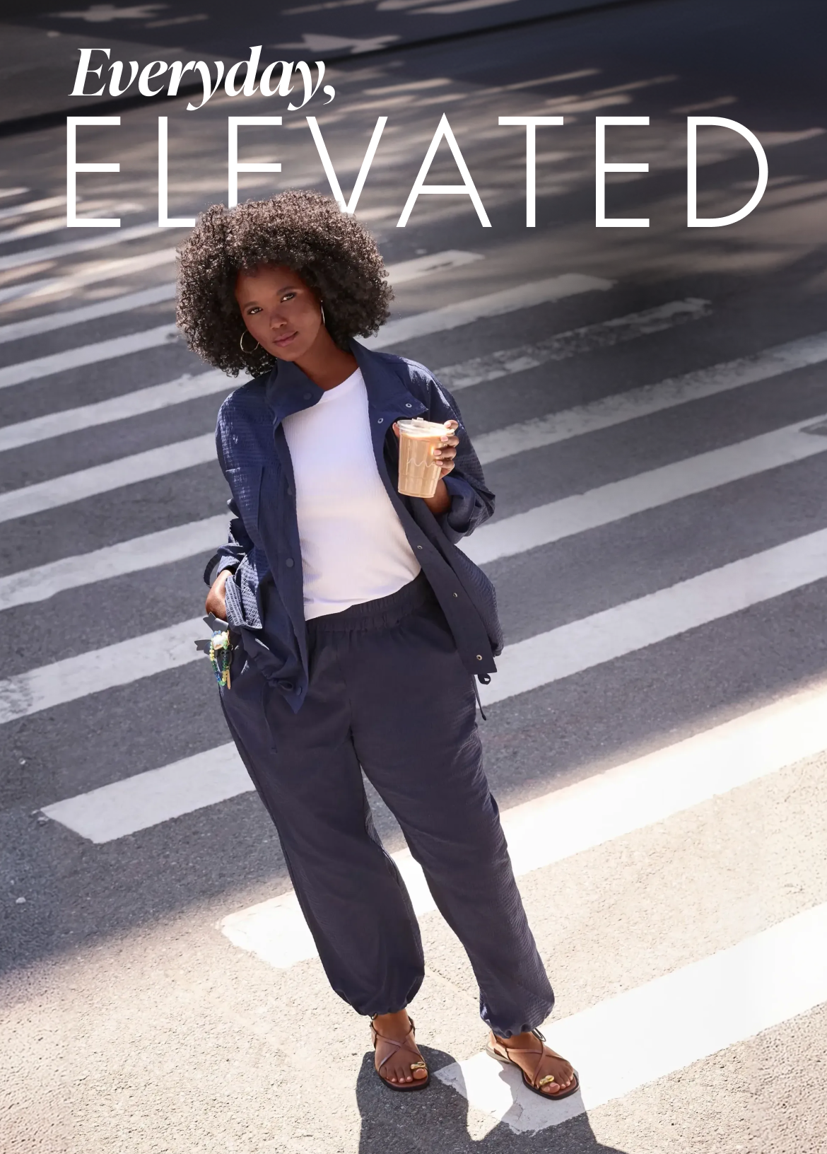 A woman with dark curly hair stands on a crosswalk holding a coffee, with the striped pattern of the crosswalk filling the background.