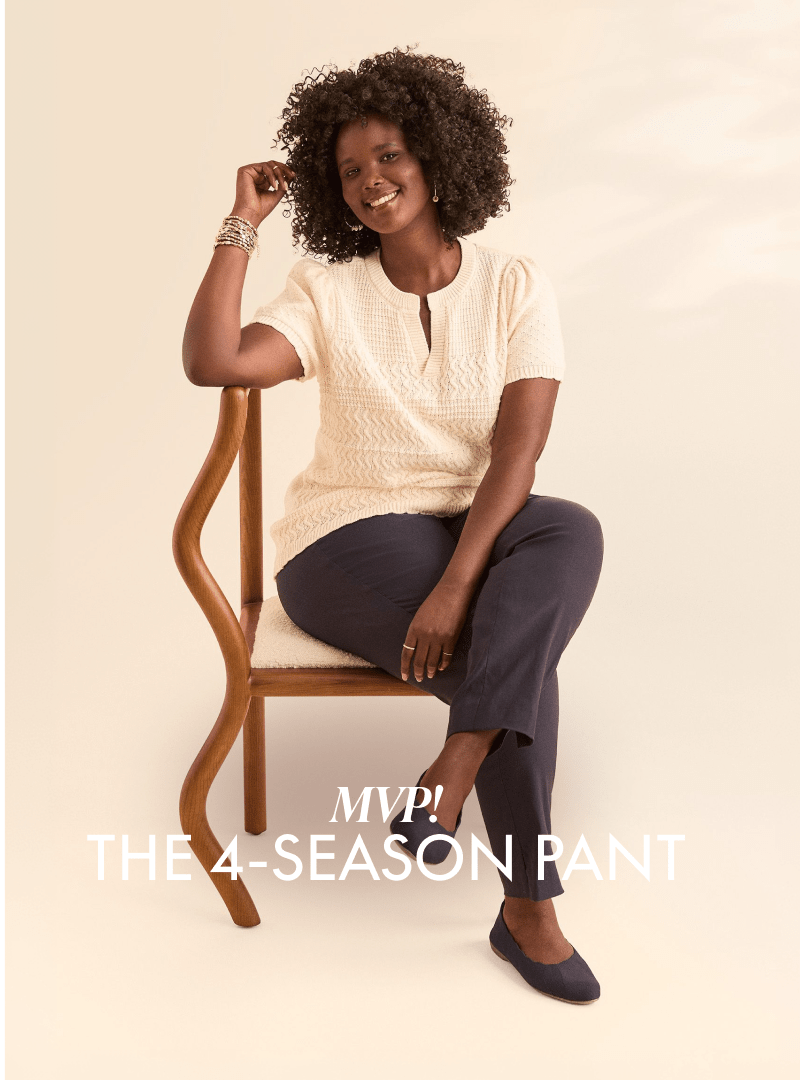 A smiling woman in a cream top and navy pants sits on a wooden chair against a plain, light background.
