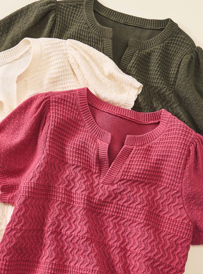 Three textured knit tops in berry, cream, and olive green are layered diagonally against a plain background.