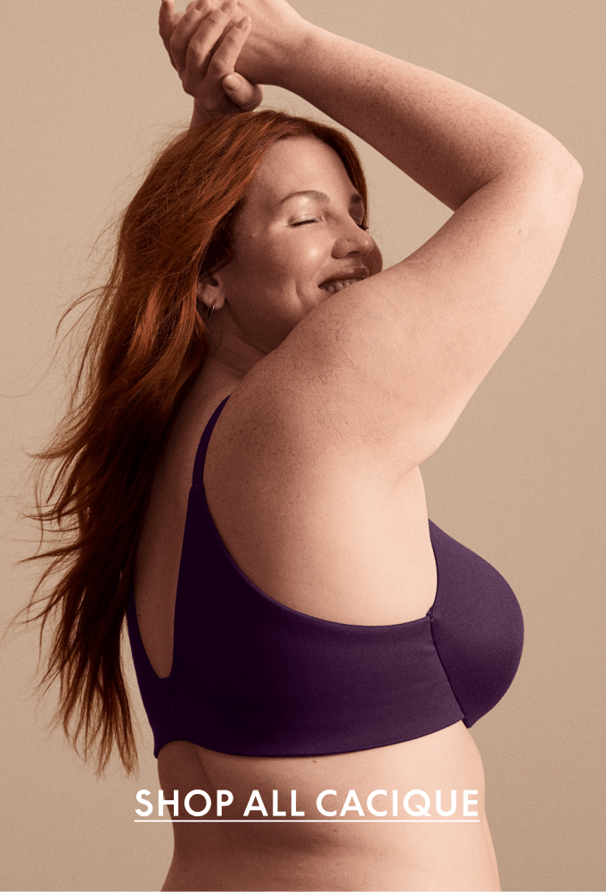 A woman with auburn hair smiles with her eyes closed, wearing a dark purple bra in the foreground, against a soft, neutral background.