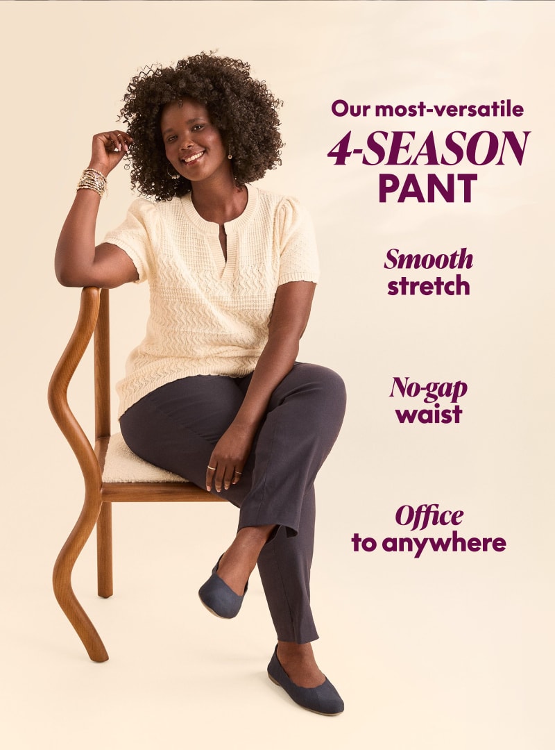 A smiling woman in a cream top and navy pants sits on a wooden chair against a plain, light background.