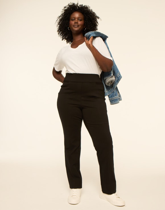 A woman in a white t-shirt, black pants, and white sneakers stands with a denim jacket slung over her shoulder against a plain, light-colored background.