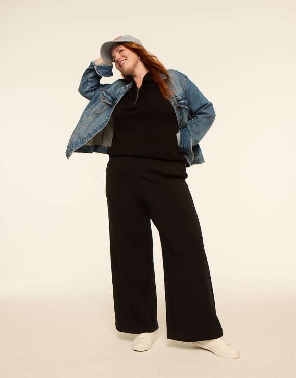 A woman in a denim jacket and black loungewear poses against a plain, light-colored background.