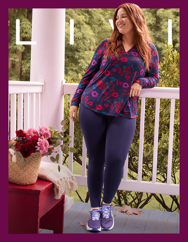 A smiling woman in a floral shirt and navy leggings stands on a porch with a basket of flowers in the foreground and trees in the background.
