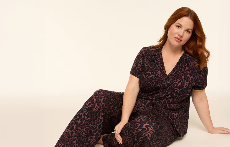 A woman with auburn hair poses in leopard-print pajamas against a plain, light-colored background.