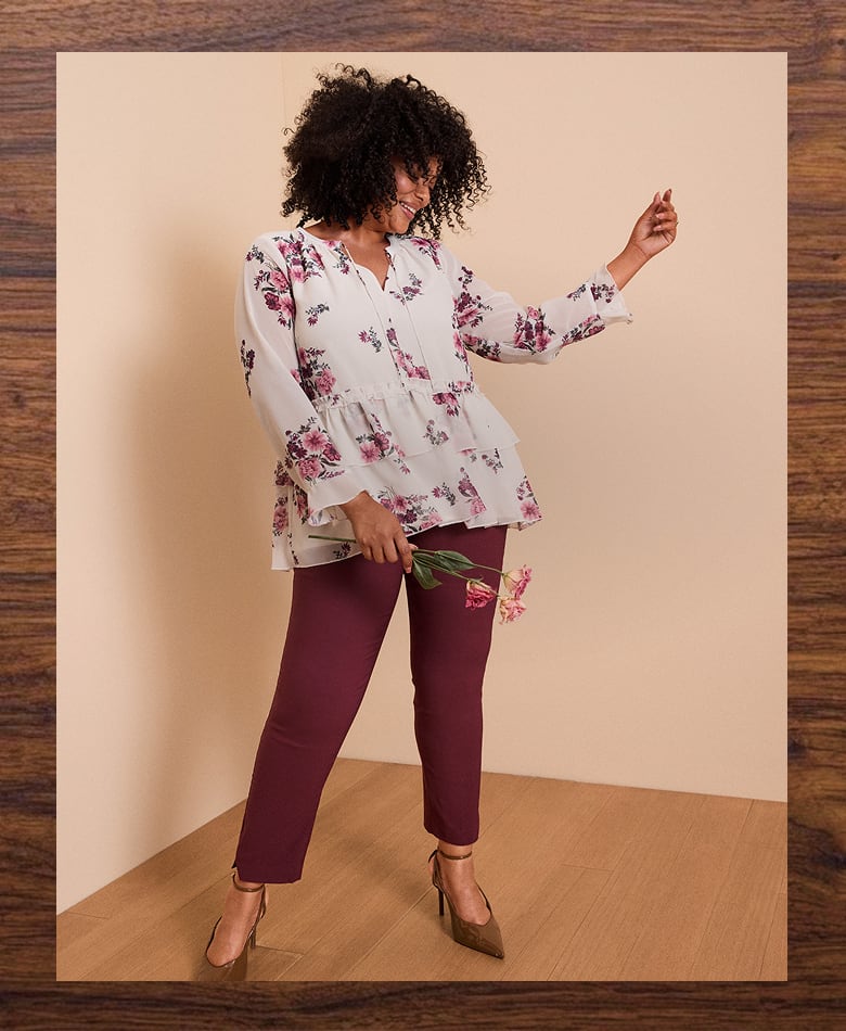A woman in a floral blouse and maroon pants poses with flowers in front of a plain background.