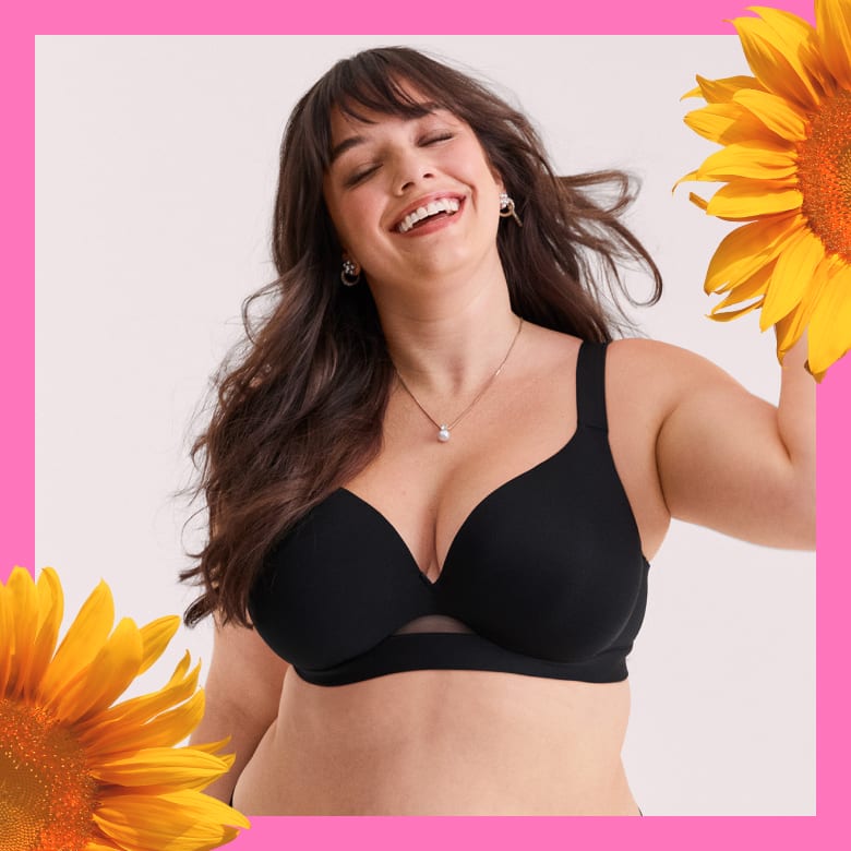 A smiling woman in a black bra is framed by sunflowers against a light pink background.