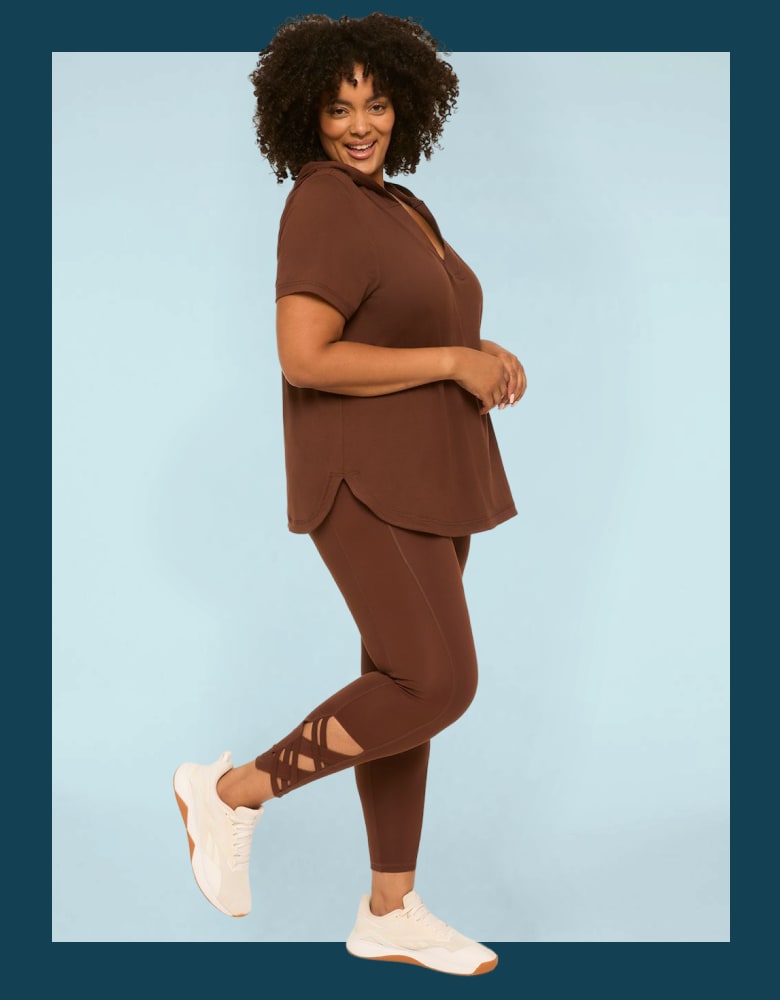 A smiling woman in a brown athleisure set with criss-cross detailing on the leggings poses against a light blue background.