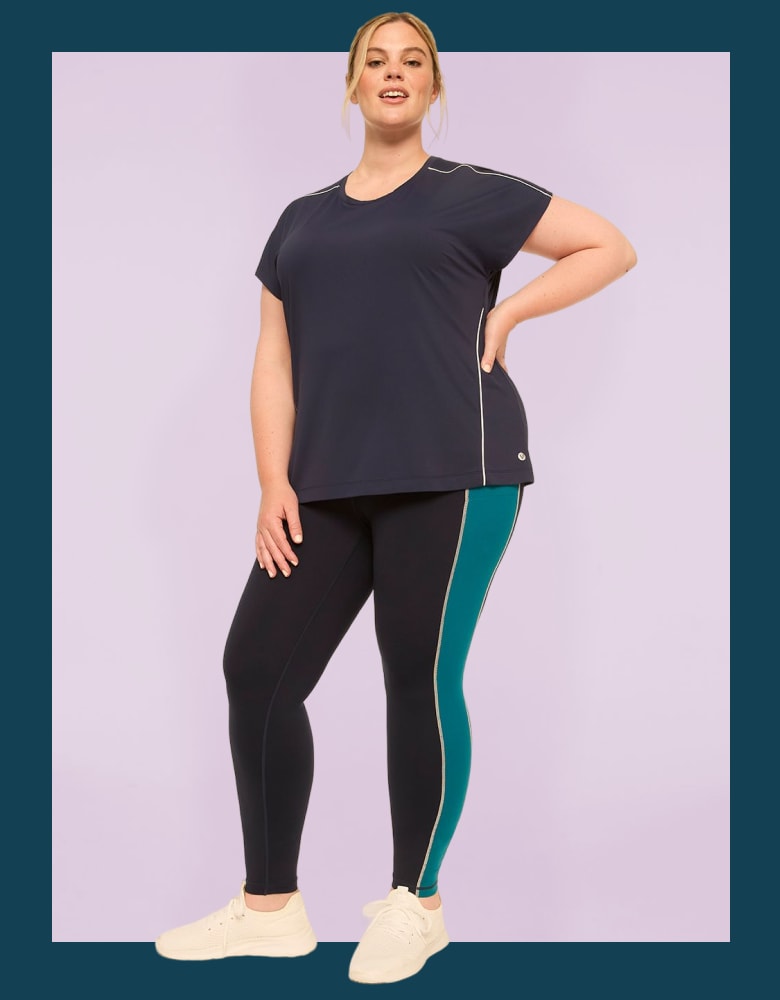 A plus-size woman in a navy t-shirt and dark leggings with a teal stripe stands against a light purple background.