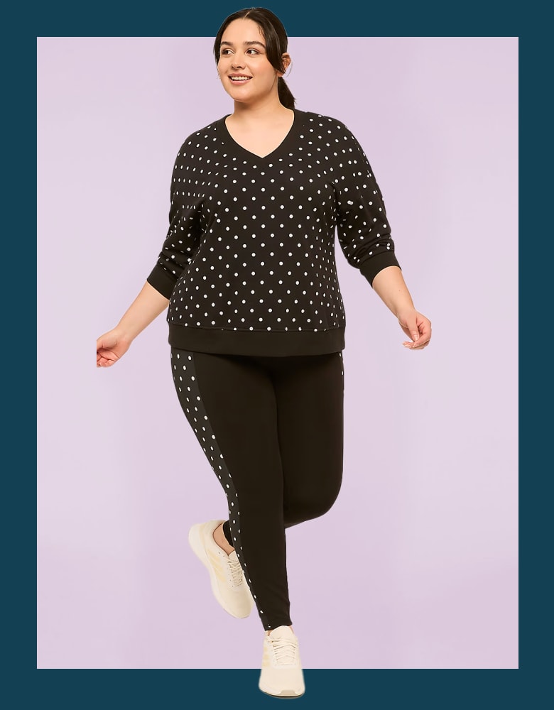 A woman in a black polka dot sweatshirt and leggings with a white stripe stands against a light purple background.
