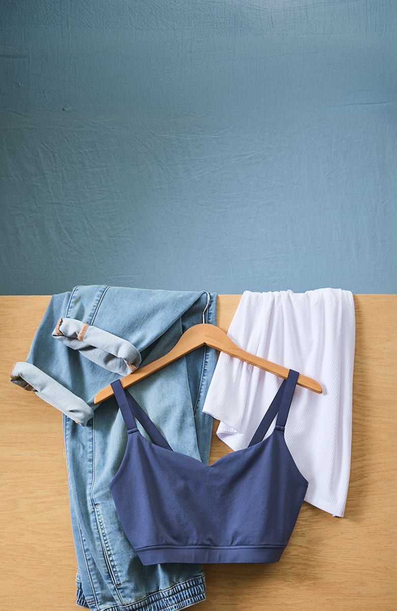 A navy blue bralette hangs on a wooden hanger between cuffed denim jeans and white shorts, set against a textured blue wall and a wooden surface.