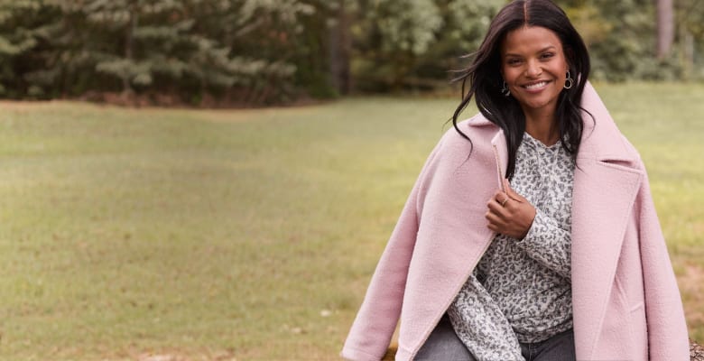 A smiling woman in a pink coat and patterned shirt sits in a grassy field with trees in the background.