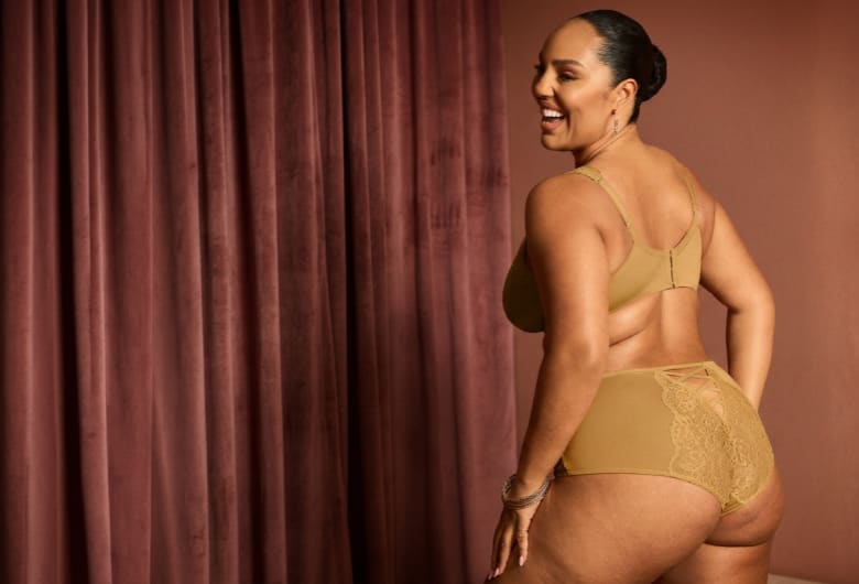 A smiling woman in tan lingerie poses in front of a draped, velvety brown curtain.