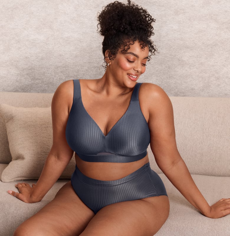 A woman in a gray ribbed bra and underwear sits on a couch with a textured wall behind her.