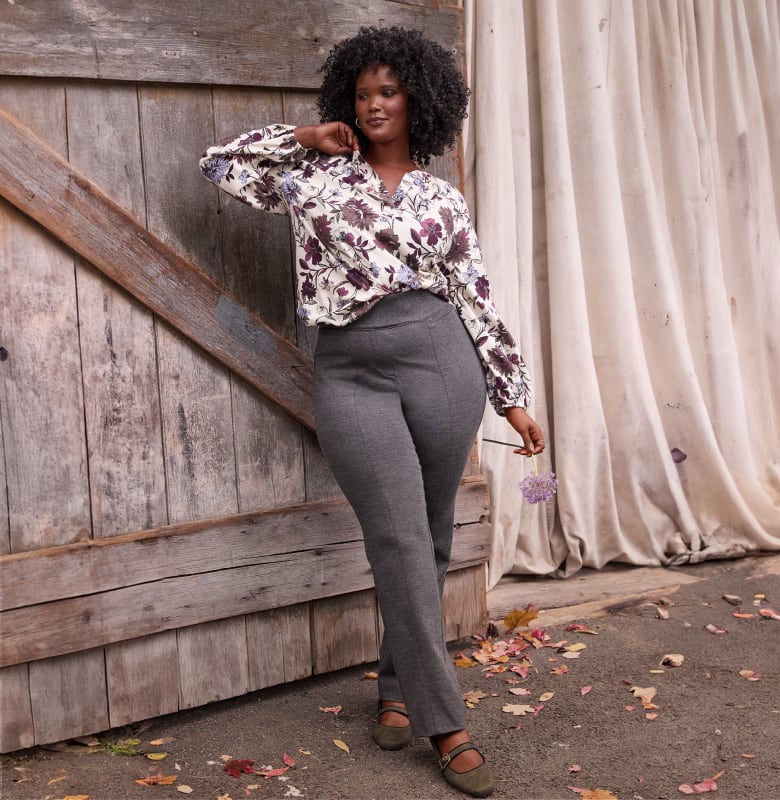 A woman in a floral blouse and gray pants poses in front of a rustic wooden door and a draped fabric backdrop.