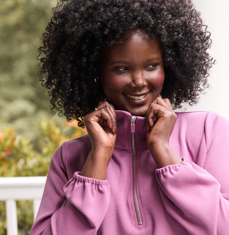A smiling woman with curly dark hair adjusts the zipper on her purple sweatshirt, with blurred green foliage in the background.