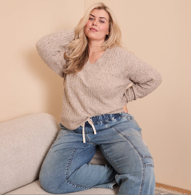 A woman in a beige sweater and blue jeans poses on a couch against a plain, light-colored wall.