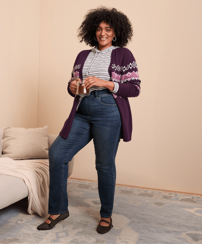 A woman in a purple cardigan and jeans stands in front of a plain wall with a couch to her left.