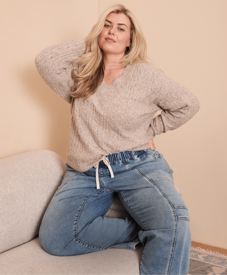 A woman in a beige sweater and denim pants poses on a couch against a peach-colored wall.