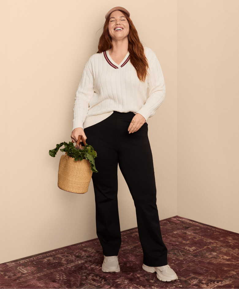 A smiling woman in a cream cable-knit sweater and black pants stands in front of a plain wall, holding a basket of greenery, with a patterned rug on the floor.