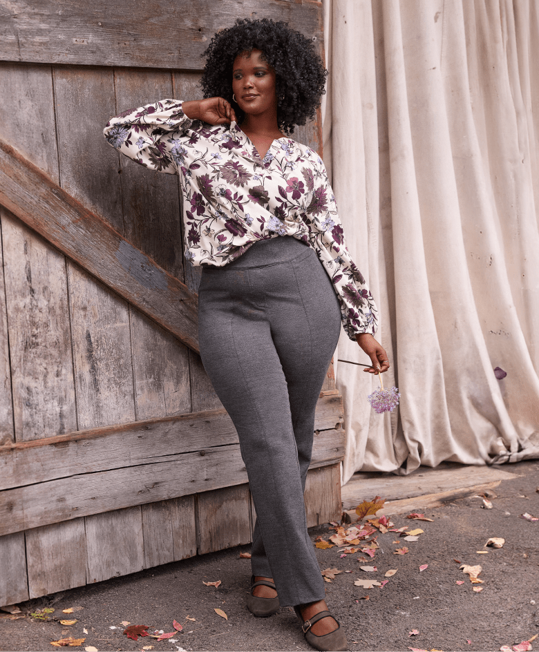 A woman in a floral blouse and gray pants stands in front of a rustic wooden door and draped fabric, with fallen leaves scattered on the ground.