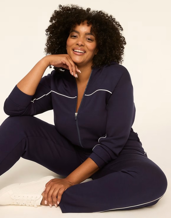 A smiling woman in a navy blue tracksuit with white piping sits on a light-colored floor against a plain, light background.