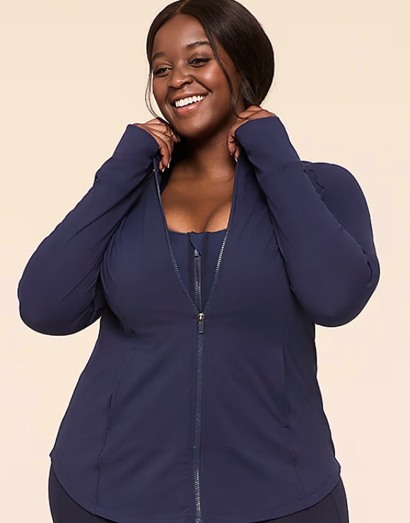 A smiling woman in a navy blue zip-up jacket and matching sports bra stands against a plain, light-colored background.