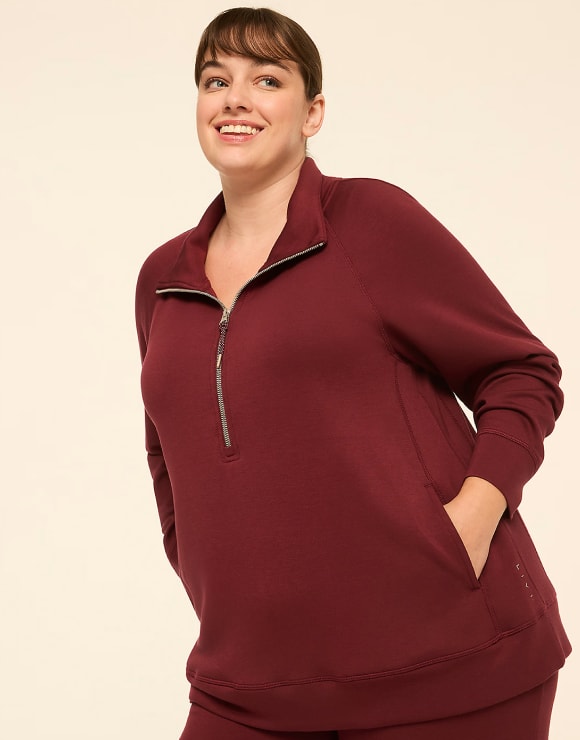 A smiling woman in a maroon quarter-zip sweatshirt with her hands in her pockets stands against a plain, light-colored background.